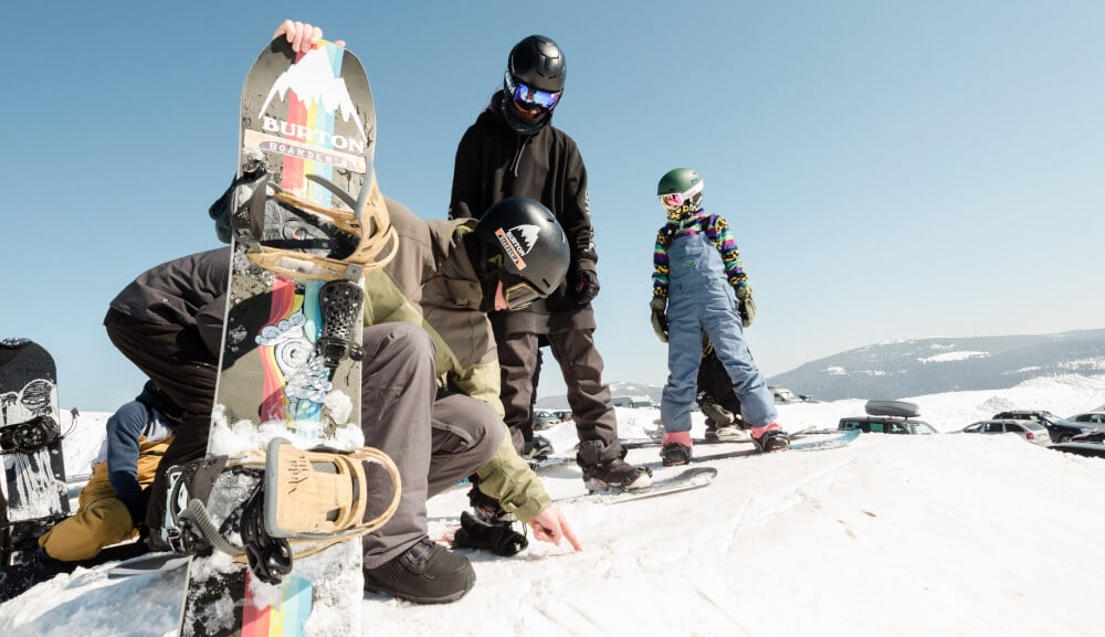 Grup de snowboarderi cu căști, ochelari și plăci, pregătind echipamentul pe zăpadă